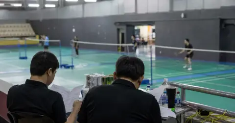 Staff review papers beside an empty badminton court as players practise