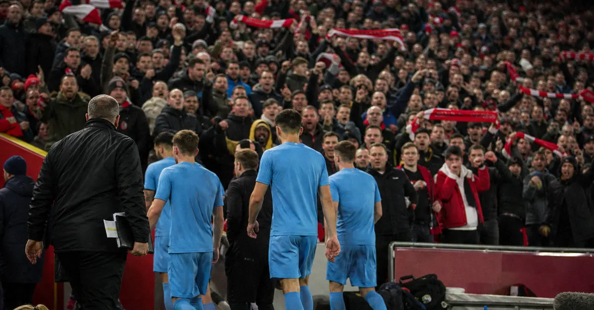 Anonymous visiting footballers face a hostile crowd beneath stadium floodlights.
