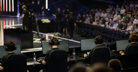 Anonymous esports players compete at computers in a crowded tournament arena.