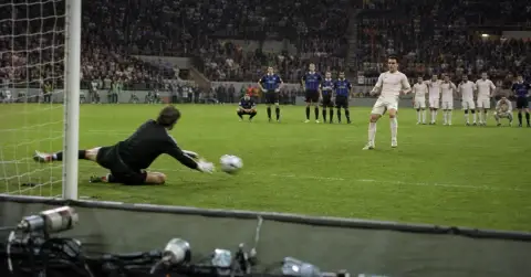 Goalkeeper dives to save a penalty as players watch from the pitch.