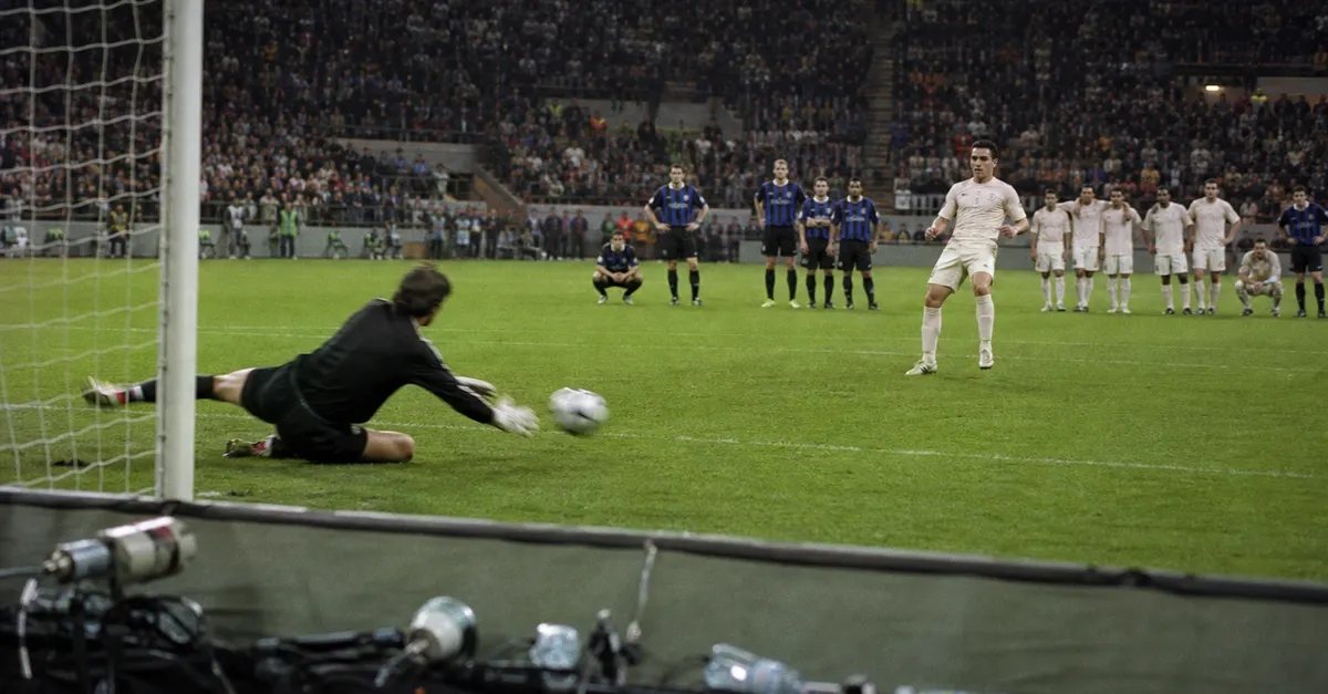 Goalkeeper dives to save a penalty as players watch from the pitch.