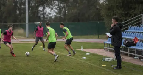 Young adult footballers train as a coach observes from the touchline.