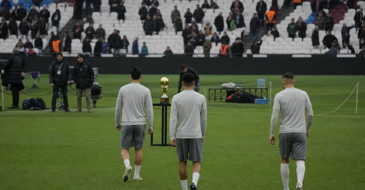 Three anonymous footballers approach a trophy at a London stadium.