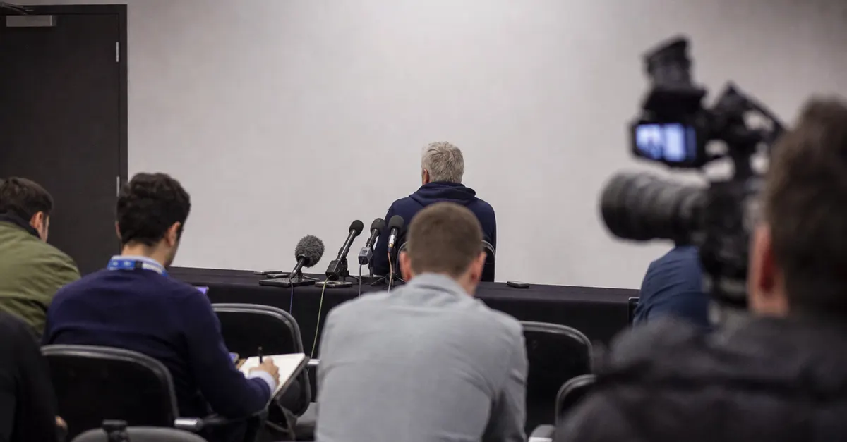 Anonymous football coach speaks at a press conference amid cameras.