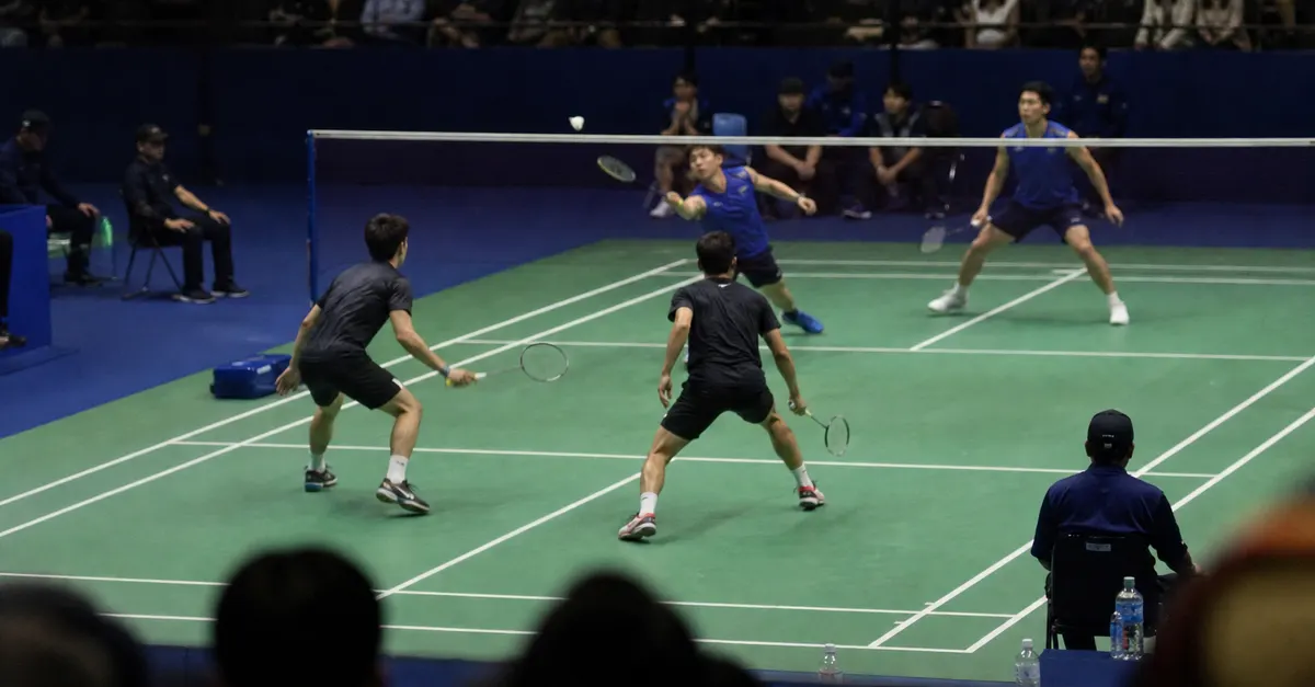 Four badminton players compete in a doubles rally inside an arena.