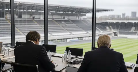 Anonymous officials review documents in a quiet football stadium boardroom.
