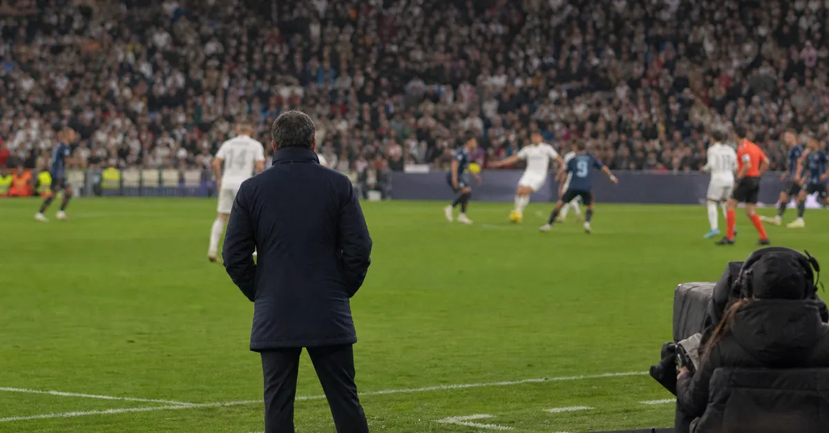 Anonymous coach watches two football sides compete in a packed stadium.