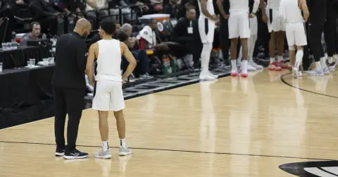 Coach speaks with a young guard beside a cluttered basketball bench