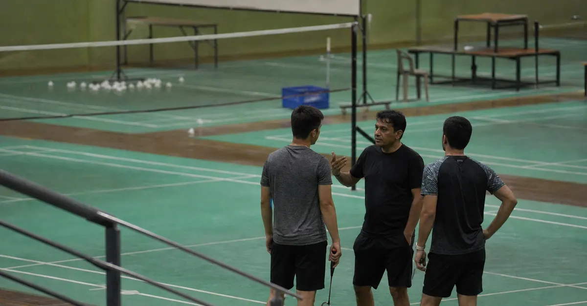 Two anonymous badminton players listen to a coach beside an indoor court.