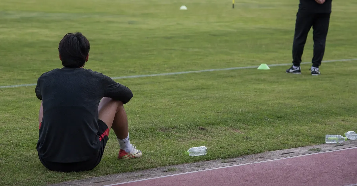 An isolated footballer sits near the touchline during a quiet training session.