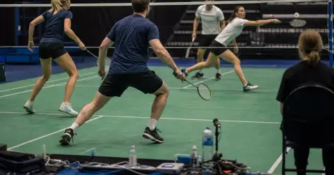 Four anonymous badminton doubles players contest a point inside an indoor arena.