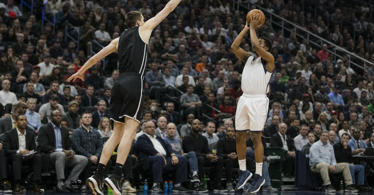 A tall defender contests a shorter guard’s jump shot during a basketball game.